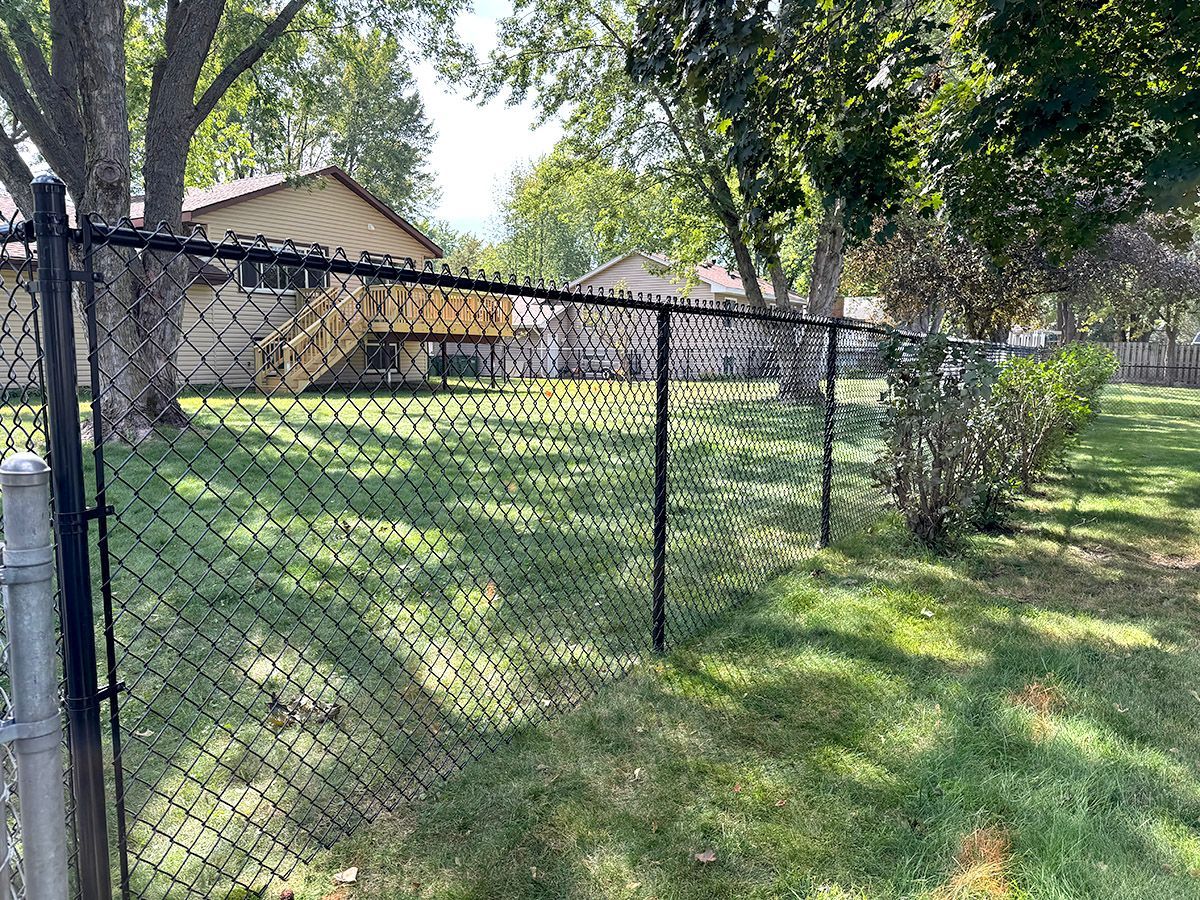 A black chain-link fence encloses a green residential backyard with trees and houses in the background on a sunny day.