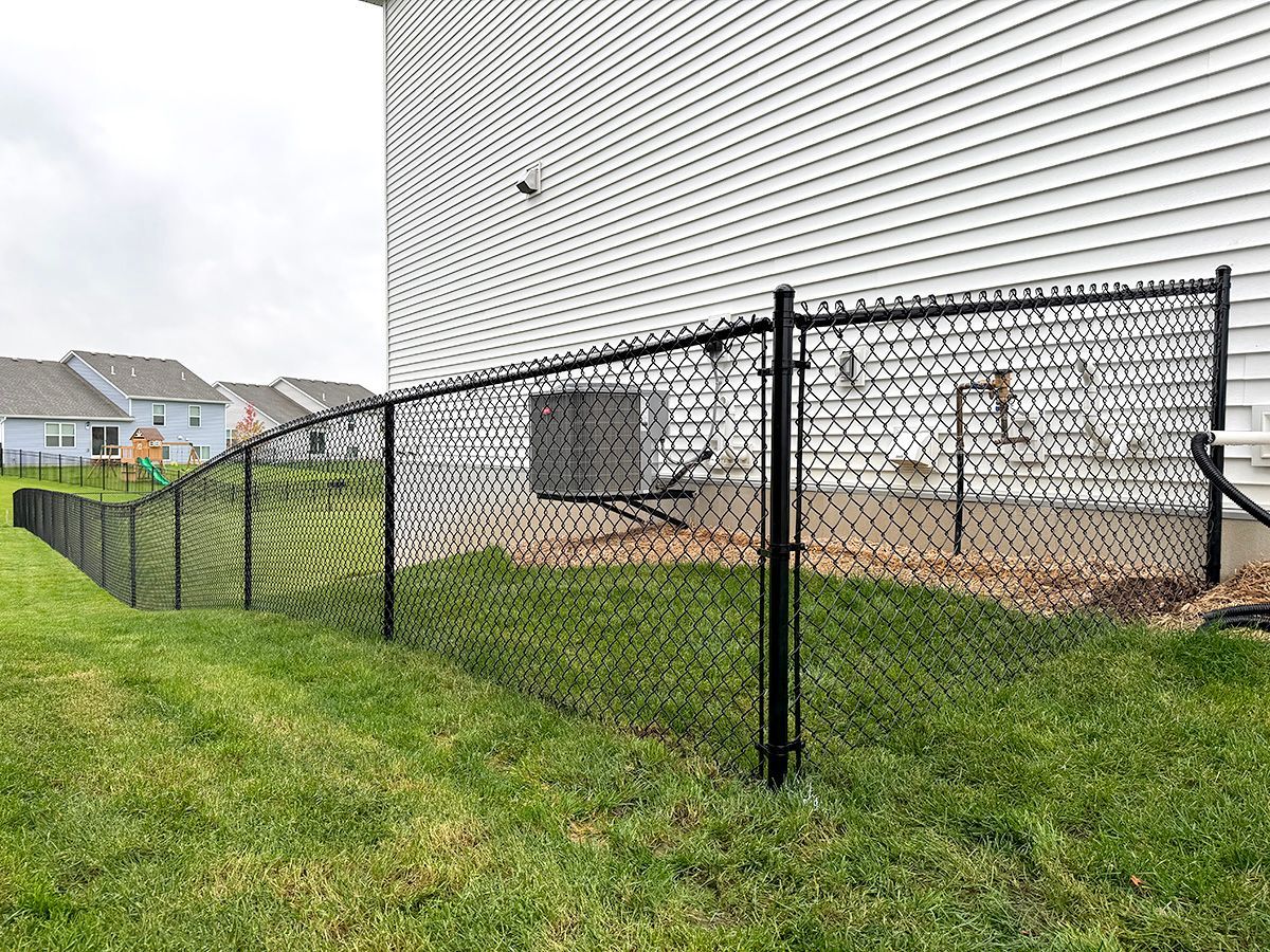 A black chain-link fence runs along the side of a white house with a green lawn and neighboring homes in the background.