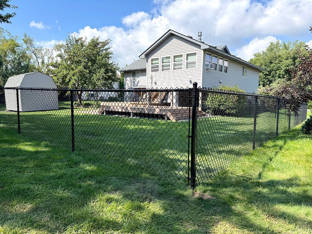 A two-story gray house with a backyard wooden deck, a storage shed, and a black chain-link fence on a sunny day.