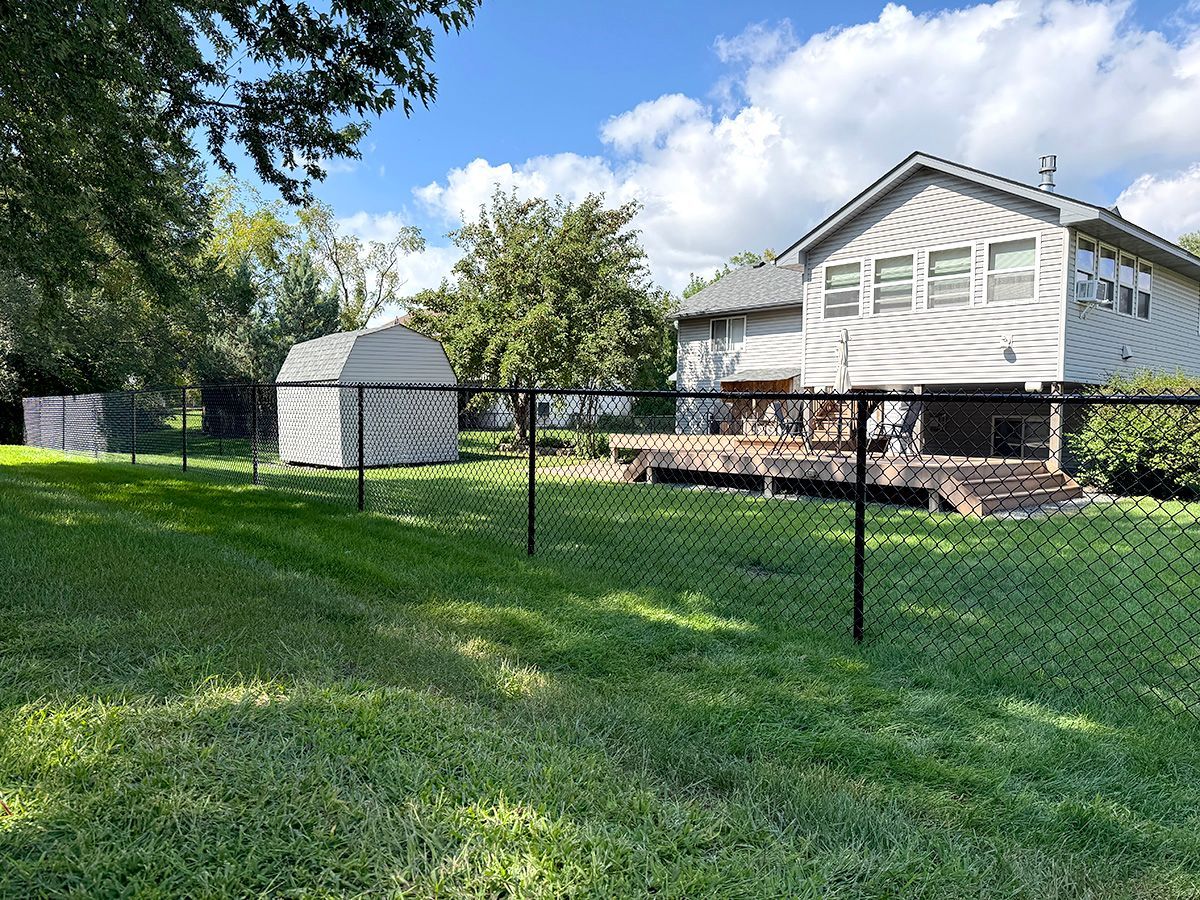 A backyard with a green lawn, a white storage shed, and a two-story house with a wooden deck, enclosed by a wire fence.