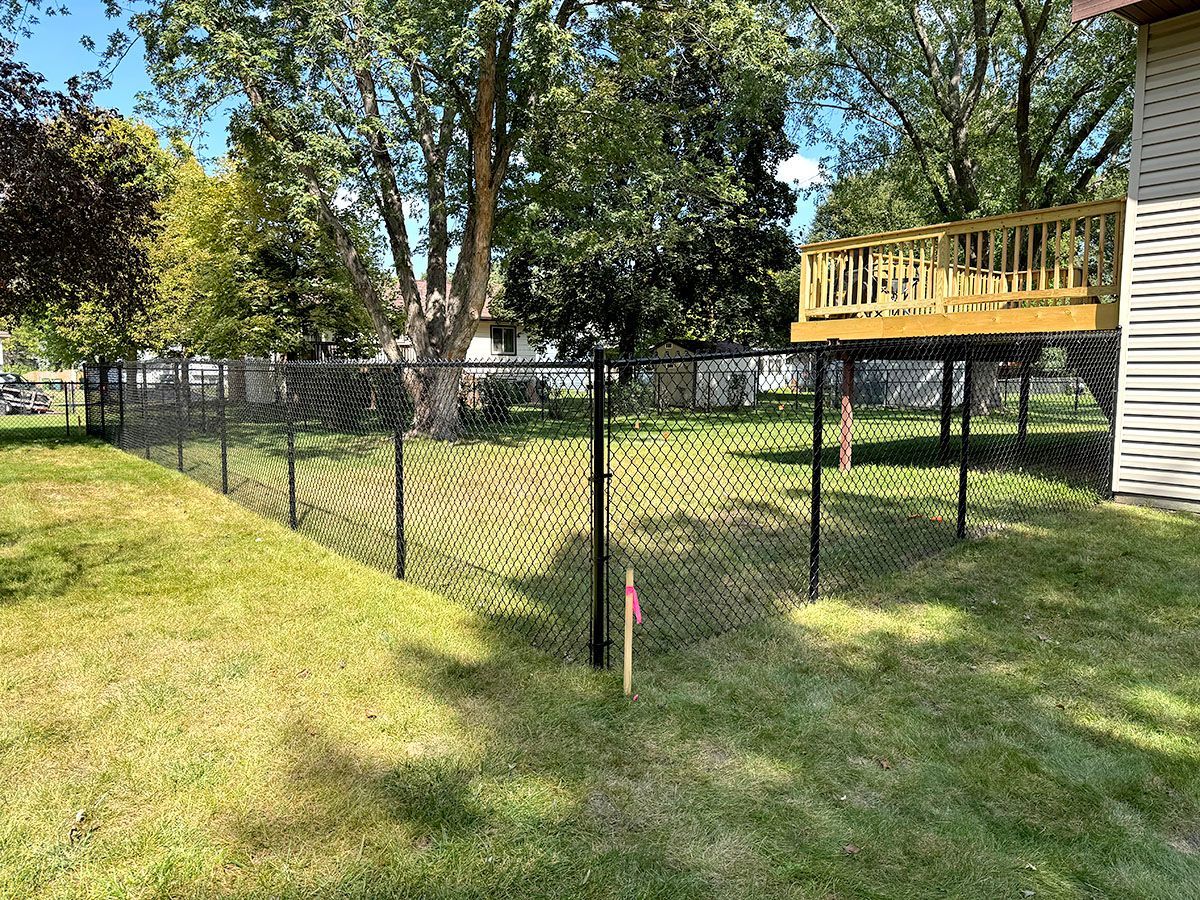 A fenced-in backyard with a black chain-link fence, green lawn, mature trees, and an elevated wooden deck on a house.