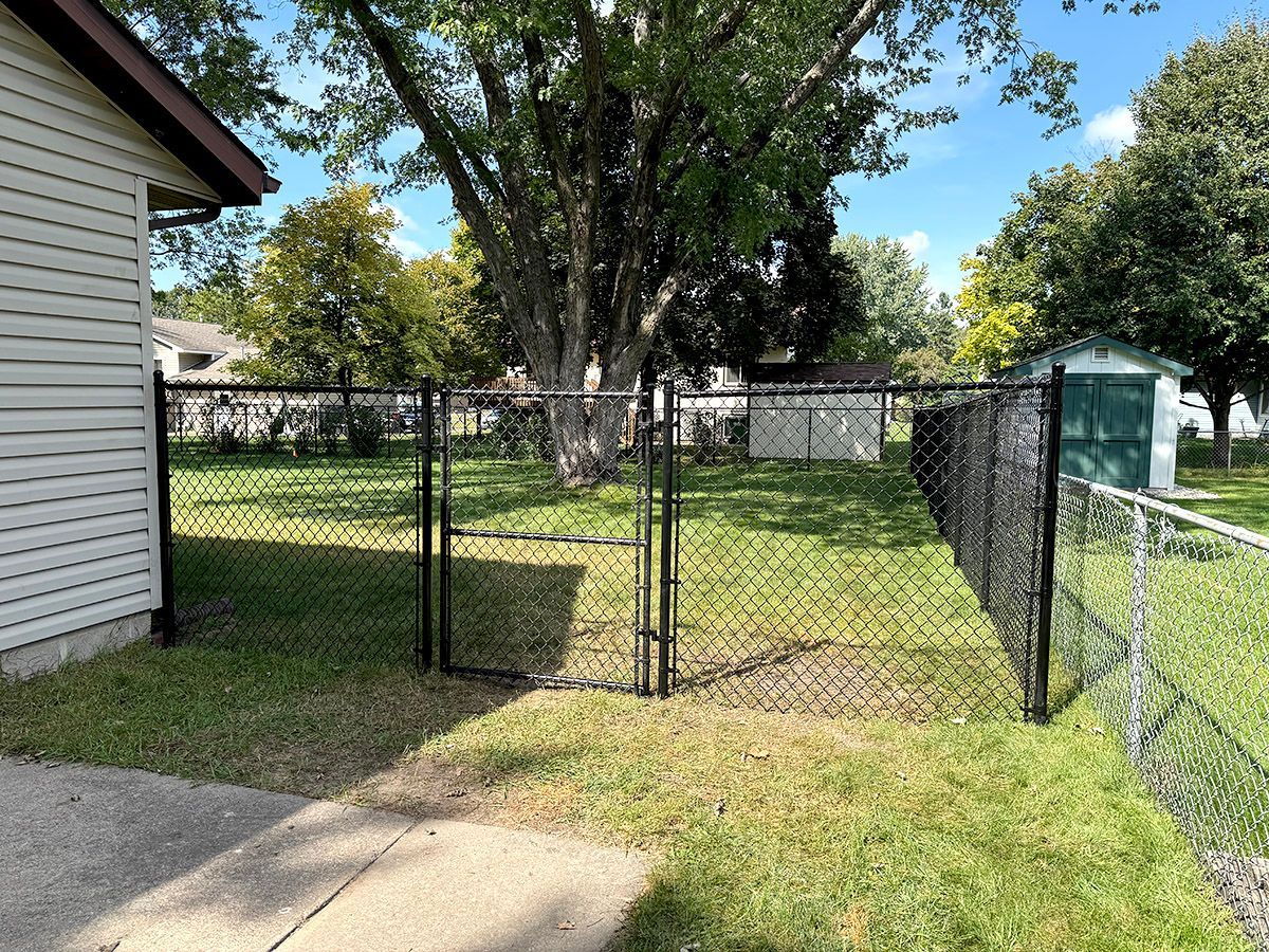 A black chain-link fence with a closed gate encloses a grassy backyard next to the white siding of a house.