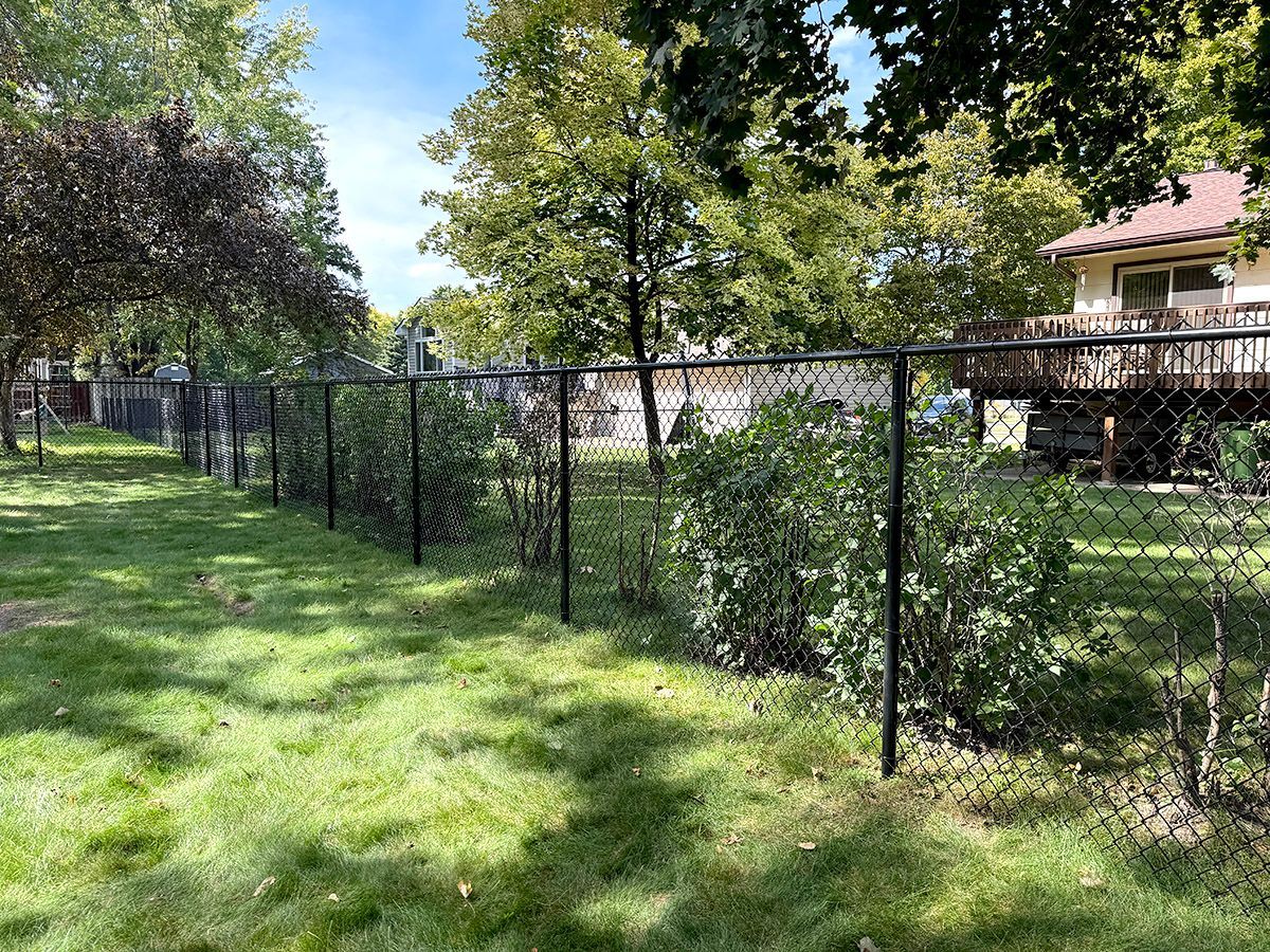 A black chain-link fence runs through a grassy backyard with several green bushes and a house with a deck in the back.