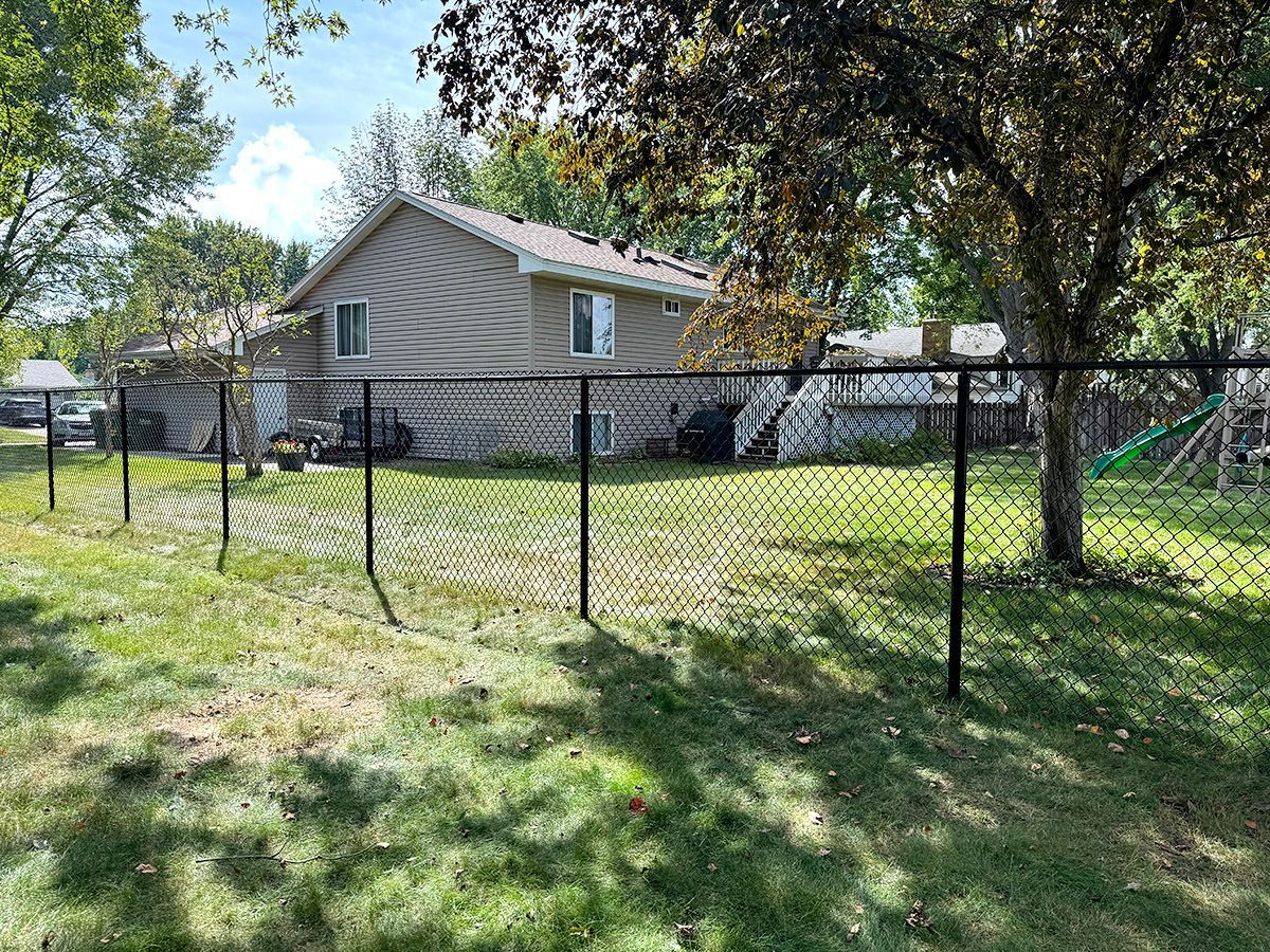 A black chain-link fence runs across a grassy yard in front of a beige two-story suburban house on a sunny day.