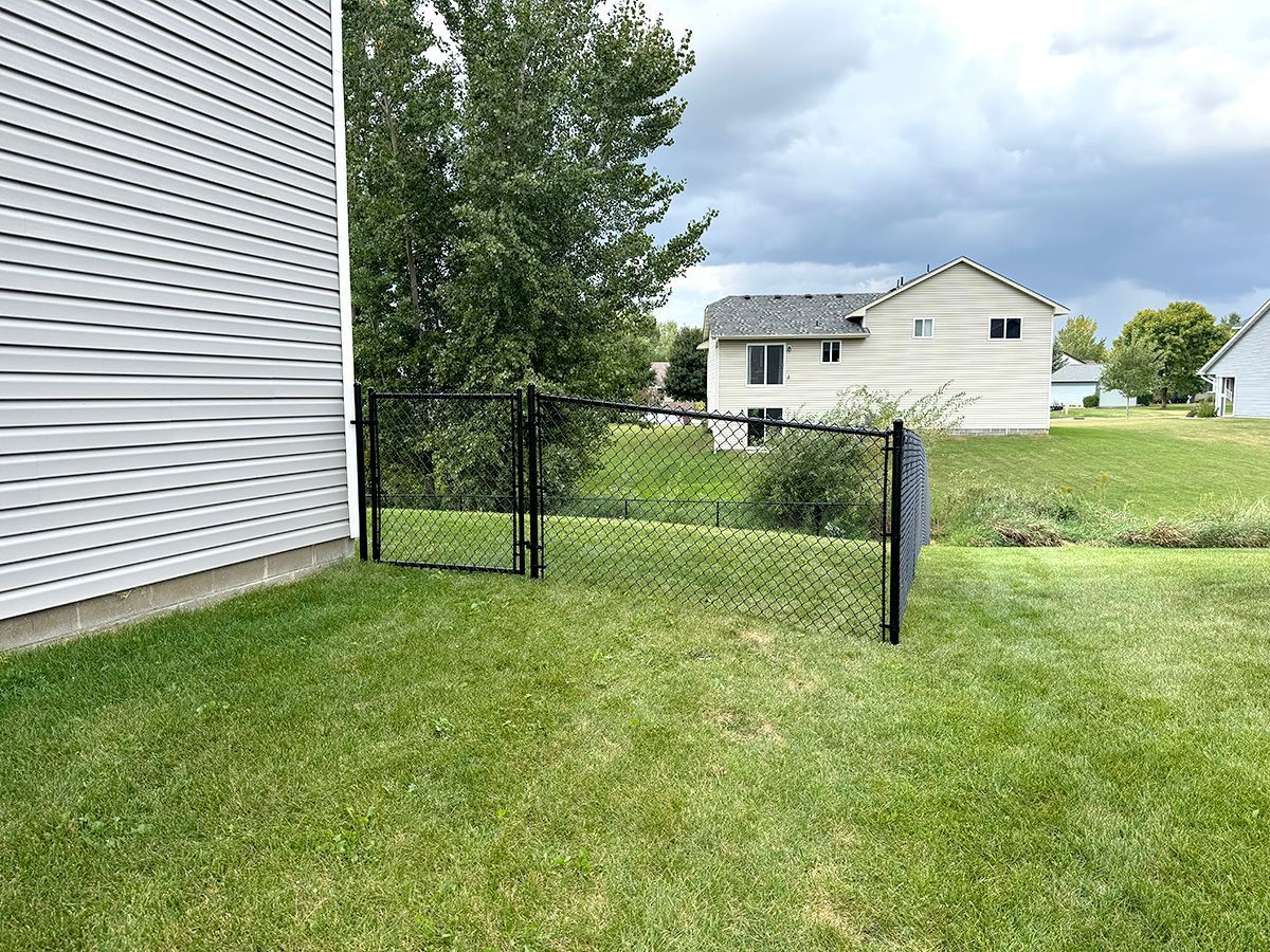 A black chain-link fence and gate extend from the side of a grey-sided house into a green backyard.