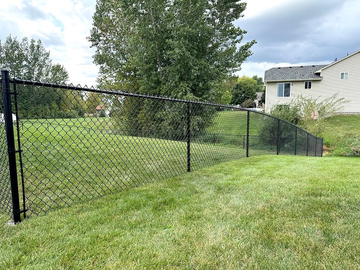 A black chain-link fence curves across a grassy backyard, with a house visible in the background under a cloudy sky.