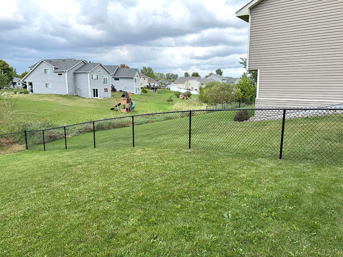 A grassy backyard slopes down toward a chain-link fence, with suburban houses in the distance under a cloudy sky.