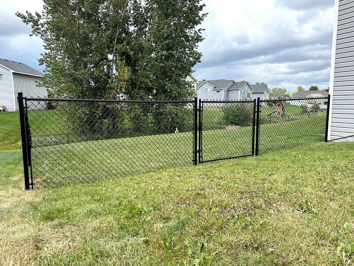 A black chain-link fence with a gate stretches across a grassy suburban backyard under a cloudy sky.