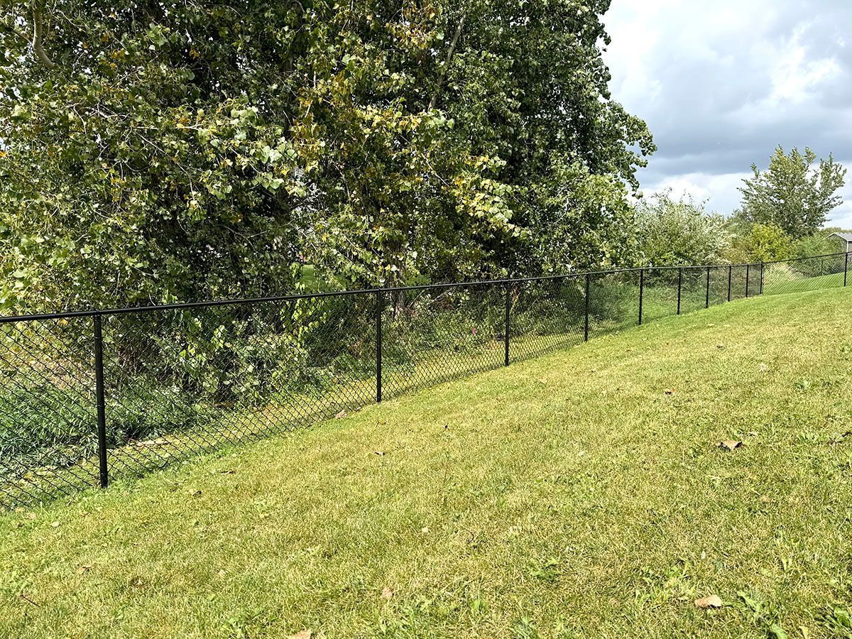 A black chain-link fence runs along a grassy, sloped hill in front of a dense line of trees under a cloudy sky.