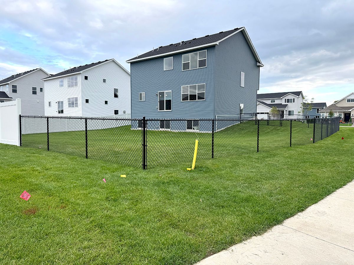 A blue two-story house with a backyard enclosed by a black chain-link fence, viewed from a suburban lawn.