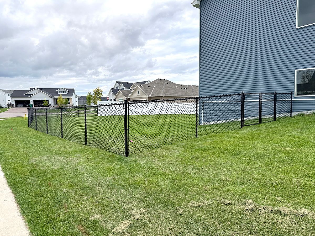 A black chain-link fence surrounds a green backyard next to a blue-sided house under a cloudy sky.