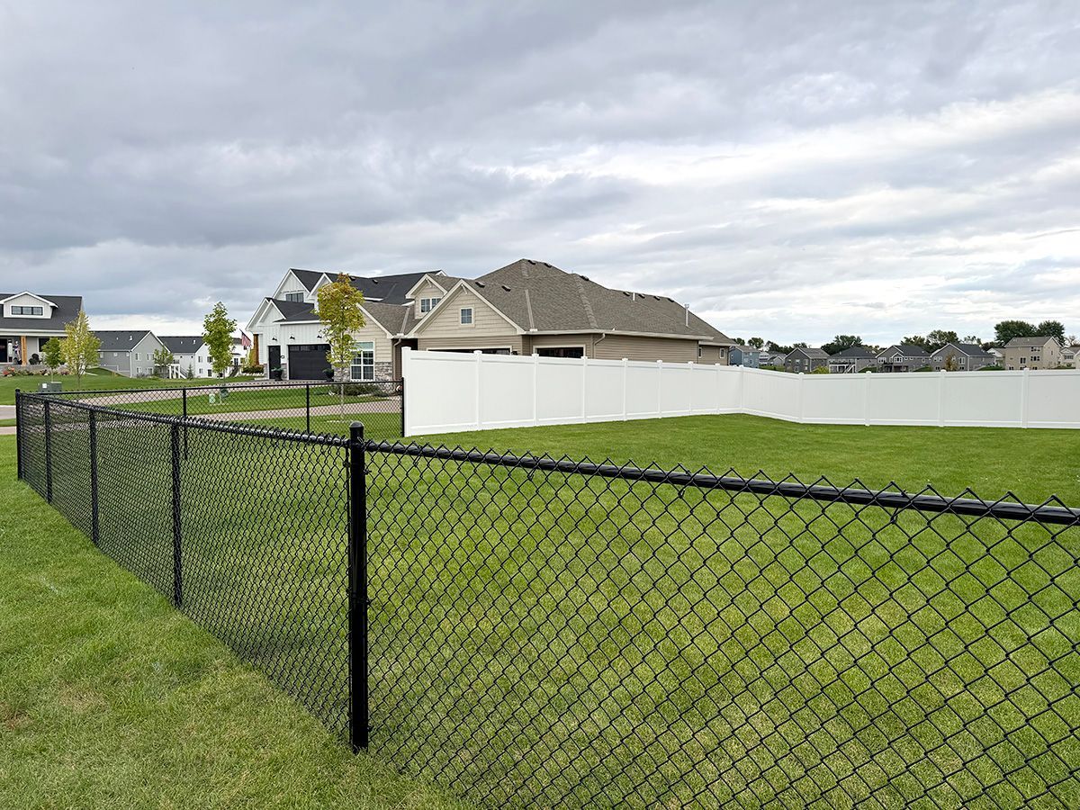 A black chain-link fence surrounds a green lawn, leading toward a house with a white privacy fence under a cloudy sky.