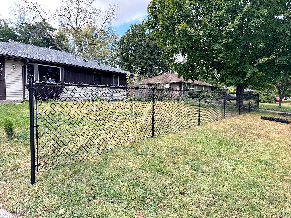 A black chain-link fence spans a grassy front yard in front of two suburban homes on a sunny day.