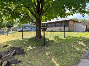 A chain-link fence surrounds a grassy front yard with a large, leafy tree and a one-story house in the background.
