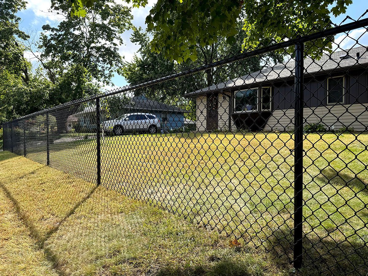 A black chain-link fence encloses a green grassy yard in front of a residential house with a car parked in the background.