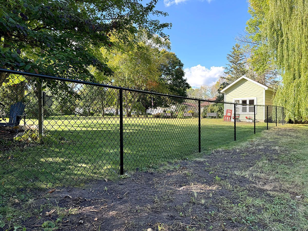 A black chain-link fence encloses a green grassy backyard with a small yellow shed and trees in the distance.
