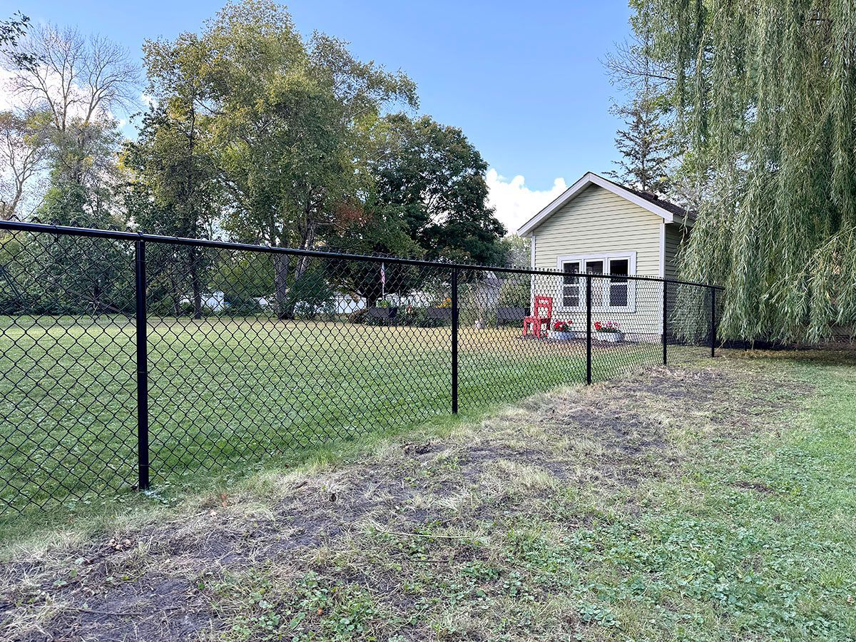 A black chain-link fence runs across a grassy lawn toward a small, light-colored house with a window and a red chair.