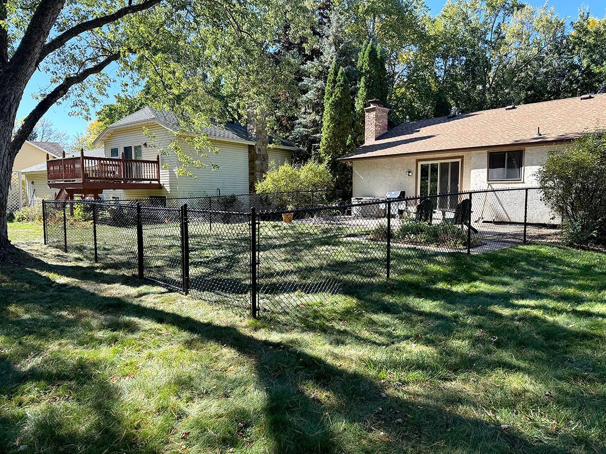 A fenced-in backyard with green grass on a sunny day, showing a house with a wooden deck and a patio area.