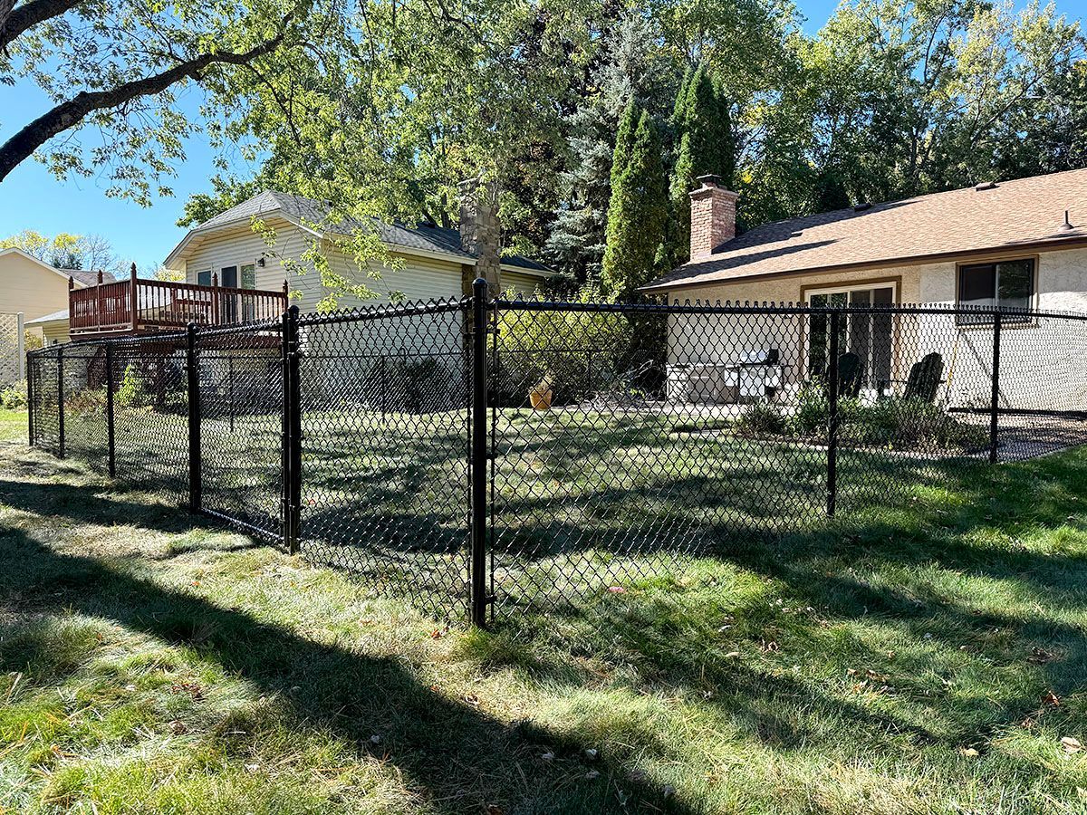A black chain-link fence encloses a green backyard lawn between two suburban houses on a sunny day.