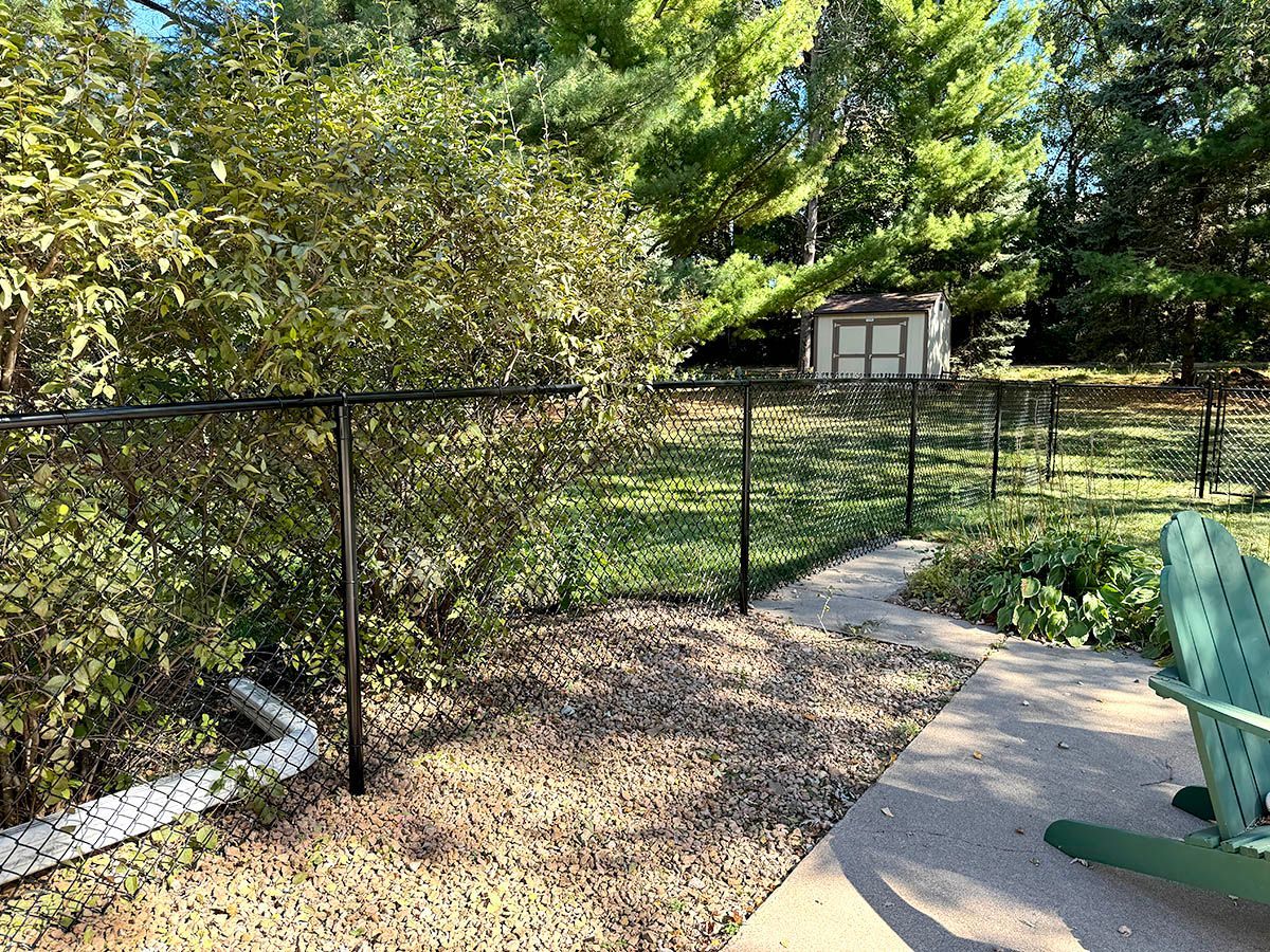 A black chain-link fence encloses a green backyard with a small shed and trees in the distance next to a stone patio.
