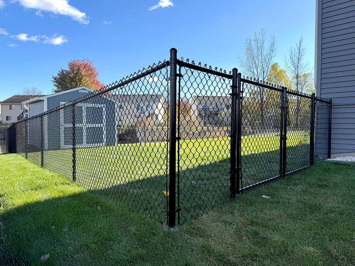 A black chain-link fence encloses a green grassy backyard with a storage shed, under a blue sky with light clouds.