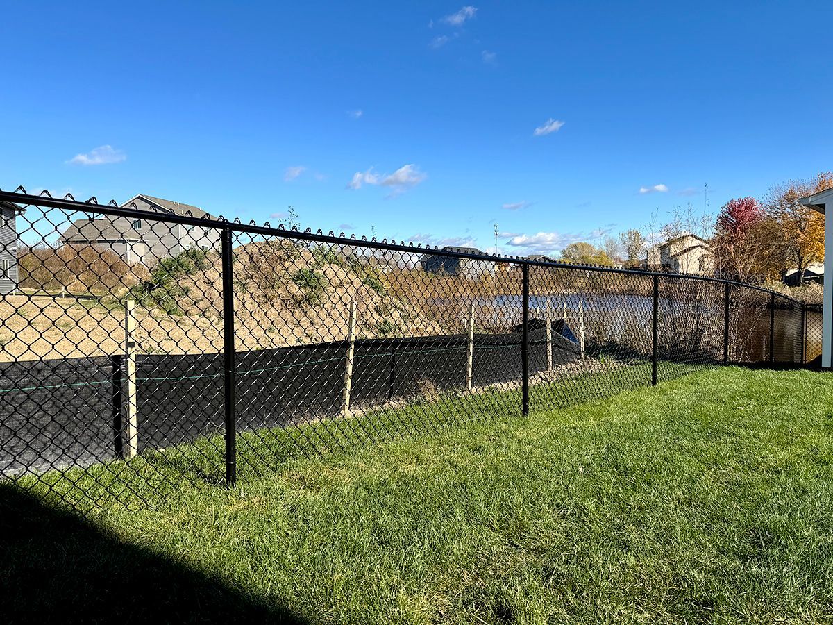 A black chain-link fence separates a green lawn from a construction area and a pond under a clear blue sky.