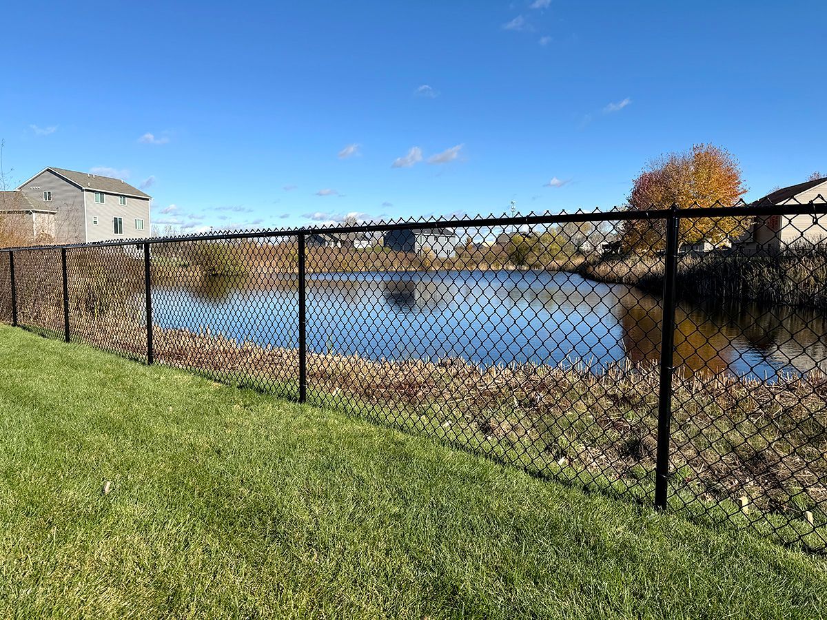 A black chain-link fence lines a green grassy lawn overlooking a calm pond under a clear blue sky.