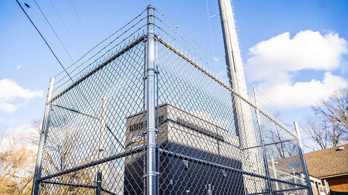 A chain-link fence topped with barbed wire surrounds a large electrical transformer box under a bright blue sky.
