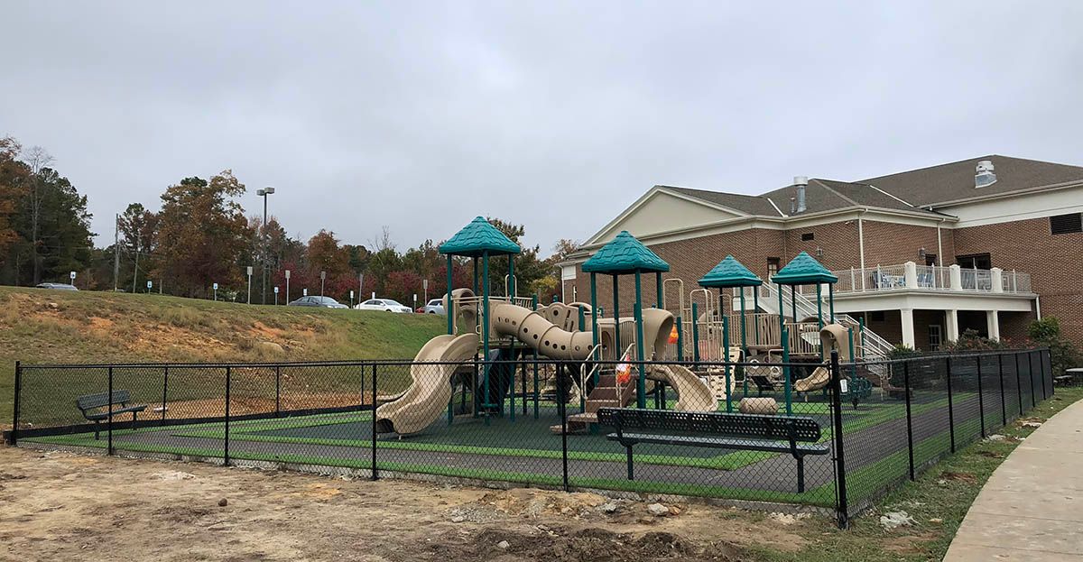 A fenced playground with slides and climbing equipment next to a brick building on a cloudy day.