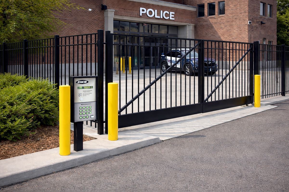 A black metal security gate blocks the entrance to a brick police station, with yellow bollards flanking a keypad.