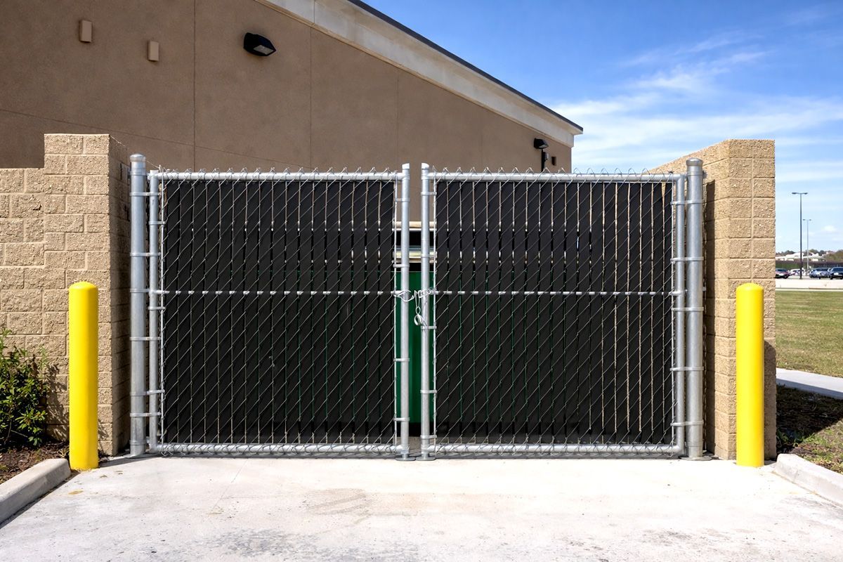 A double chain-link gate with black privacy slats, flanked by two yellow bollards and concrete block walls.