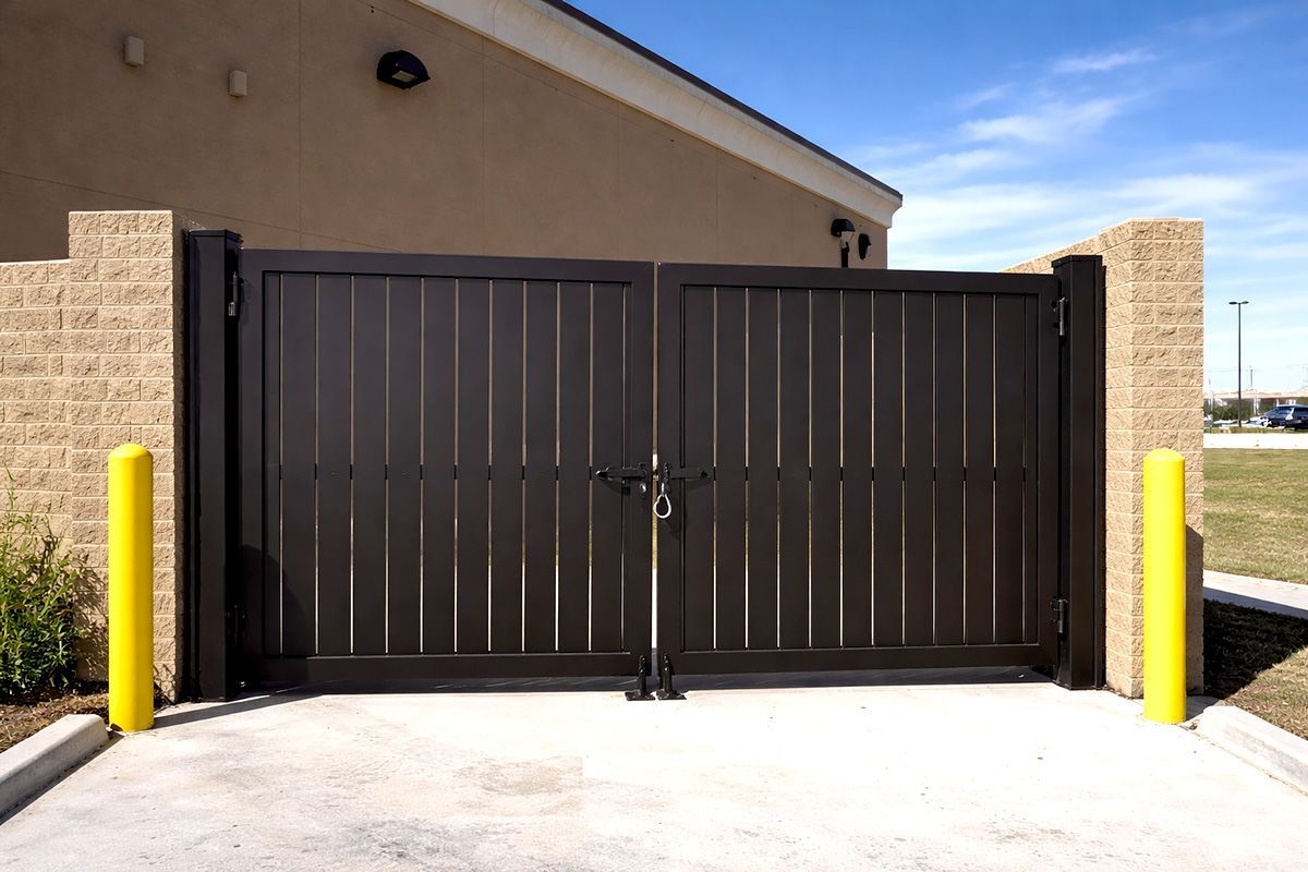 A dark brown, double-swing metal gate with vertical slats, situated between two tan brick pillars and yellow bollards.