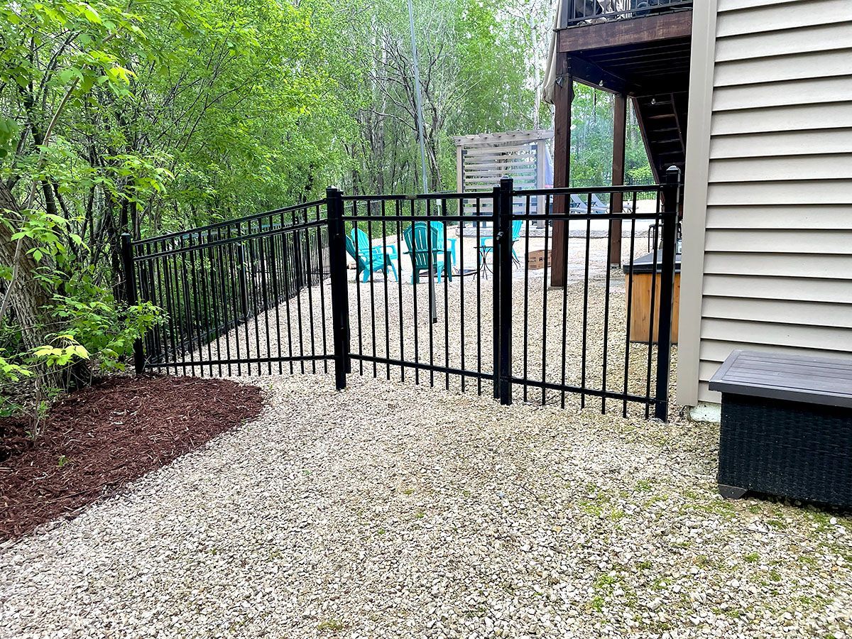 A black metal fence encloses a gravel backyard patio area with teal chairs next to a house with beige siding.