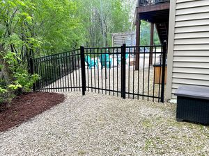 A black metal fence enclosure on a gravel patio beside a house, featuring two teal Adirondack chairs and lush greenery.