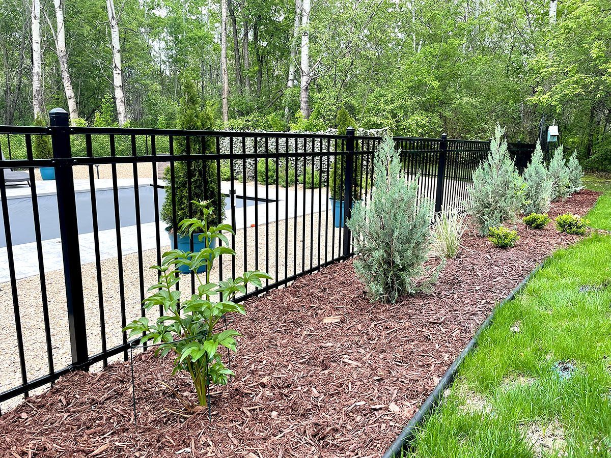 Black metal fence beside mulched shrubs and a green lawn along a wooded roadside