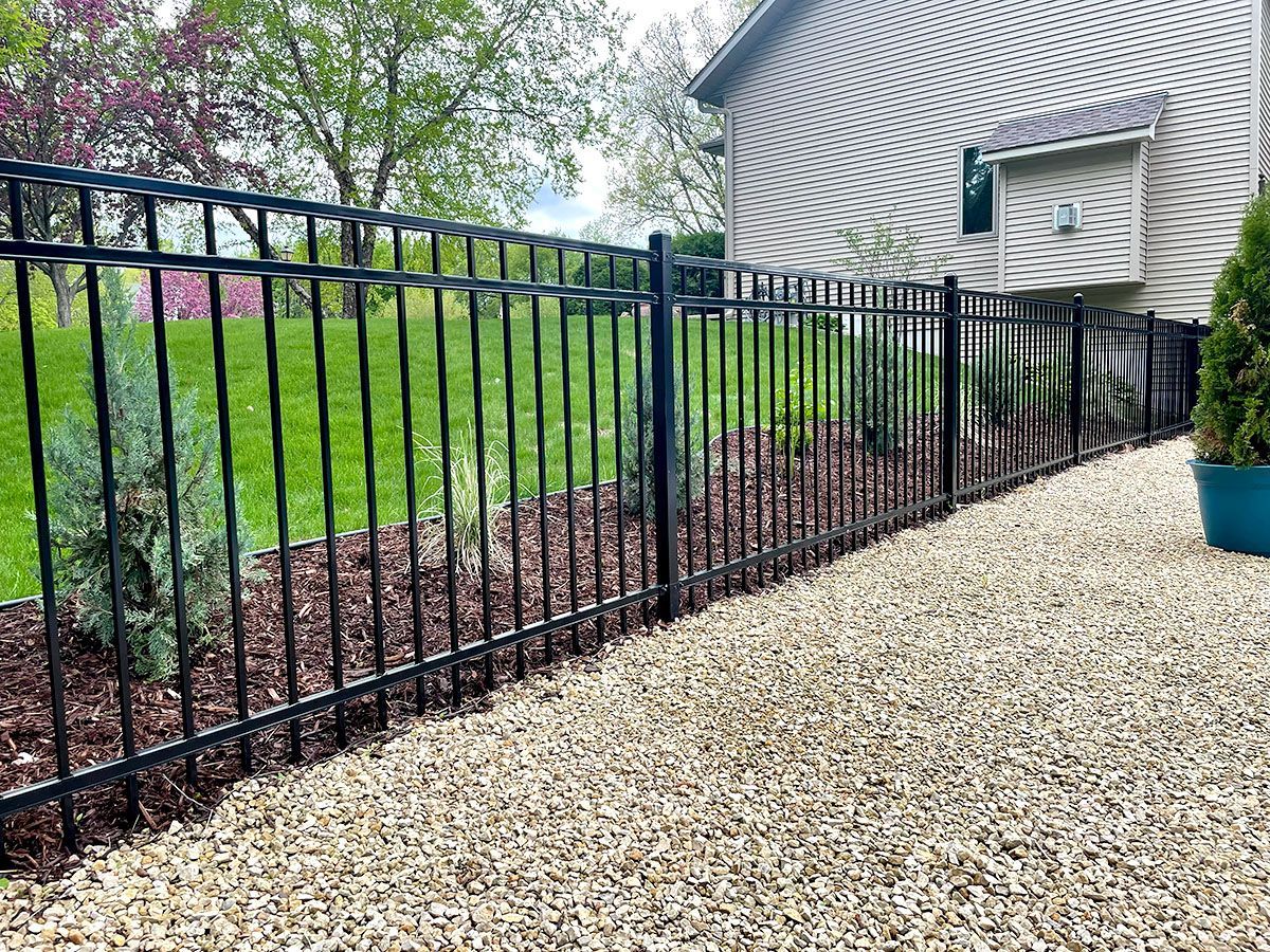 A black aluminum fence borders a gravel path, a mulched garden bed with small evergreen shrubs, and a beige house.