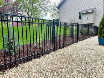 A black metal fence runs along a gravel path next to a residential house with mulch and small evergreen trees.