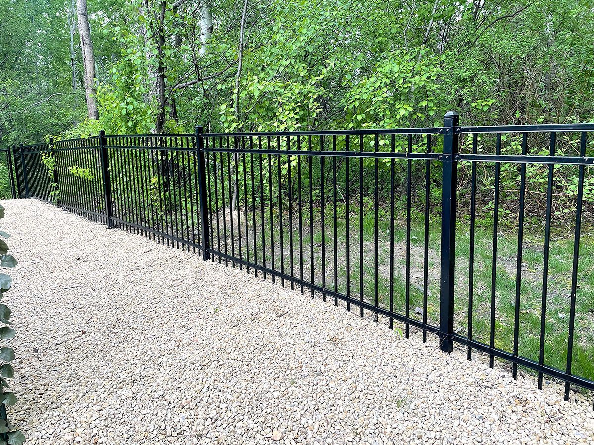 A black metal picket fence runs along a gravel path in front of a backdrop of green trees.