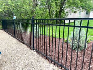 A black metal picket fence separates a gravel path from a garden bed with mulch and shrubs, backed by green trees.