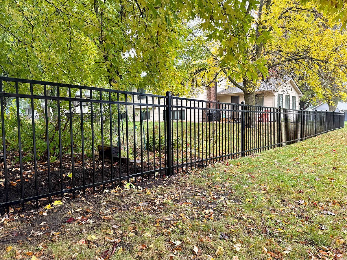 Black metal fence along a leafy yard beside a house under green autumn trees.
