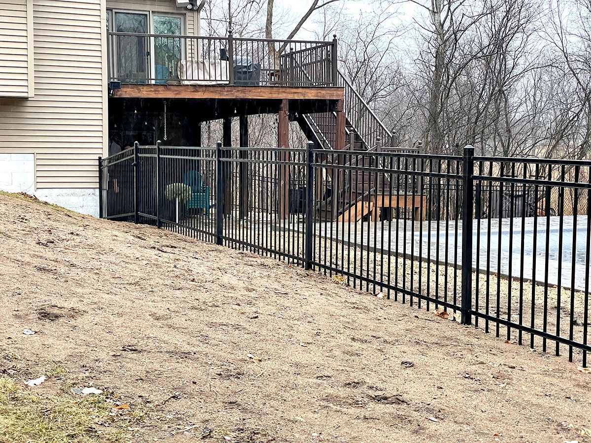 A metal fence separates a grassy, sloping yard from a house with a raised wooden deck in the background.