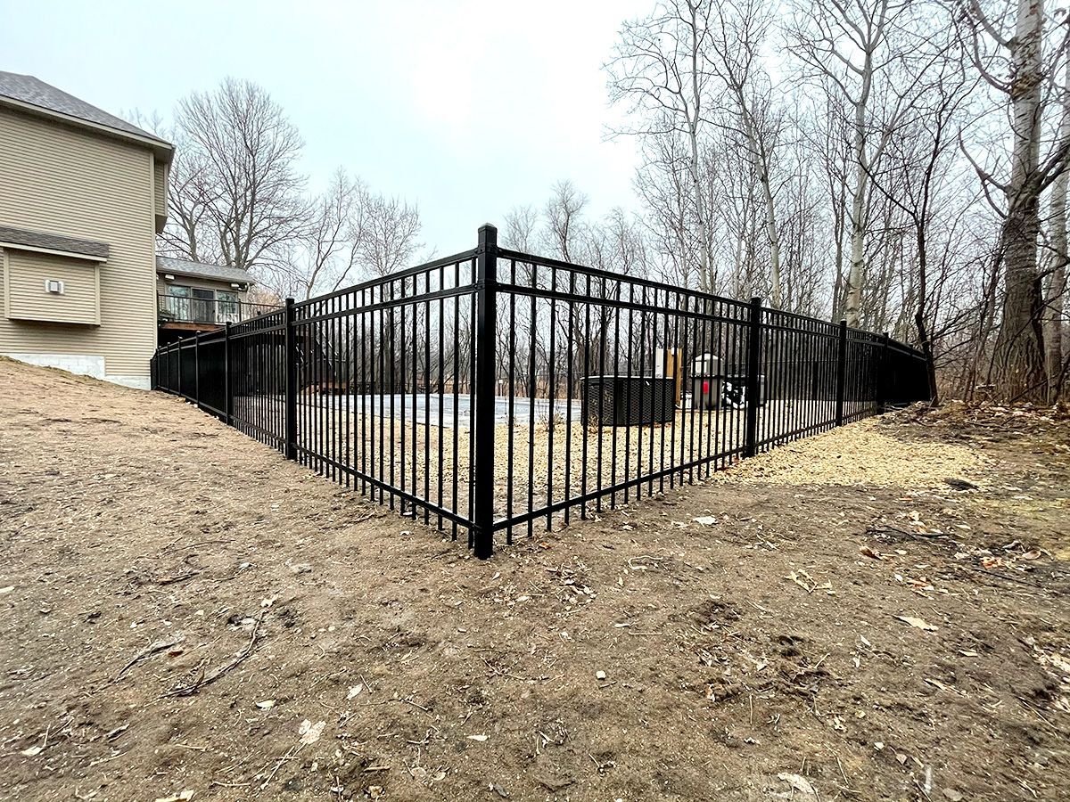 A black metal fence encloses a backyard pool area on a cloudy day, set against a backdrop of bare trees and a house.