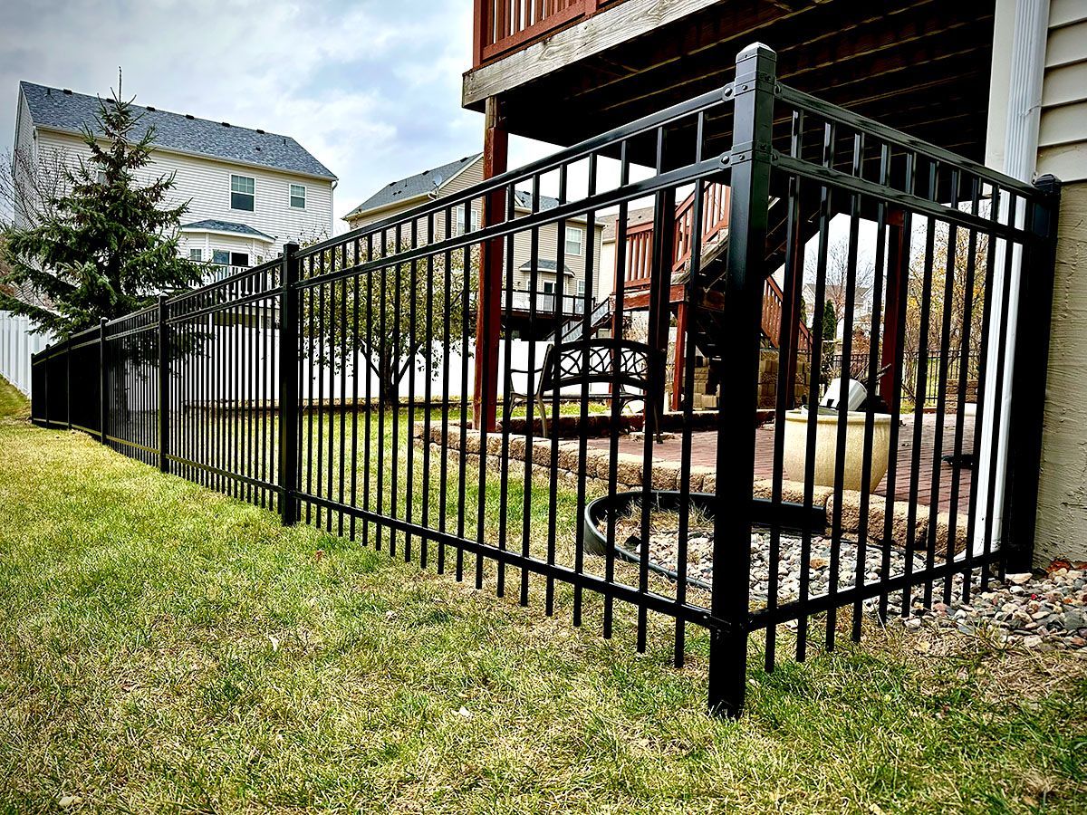 A black aluminum fence runs along a green residential lawn next to a house with a wooden deck.
