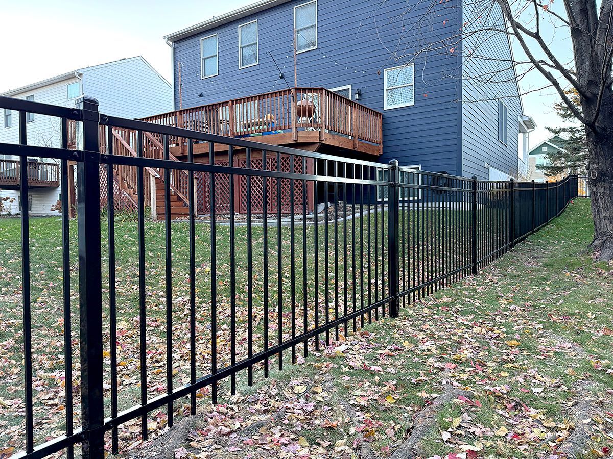 A black metal picket fence runs through a grassy backyard with fallen leaves, set against a blue house with a wood deck.
