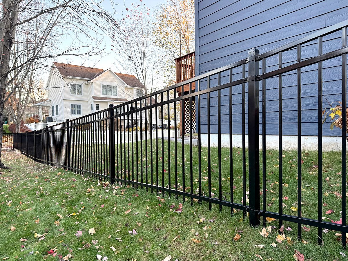A black metal fence runs along a sloping grassy lawn next to a blue house, with a white suburban home in the background.