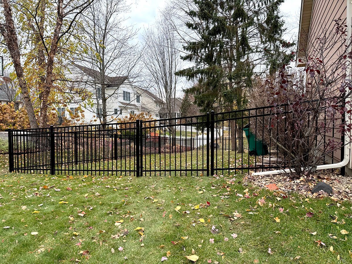 A black metal fence runs across a residential lawn near a house, with bare trees and neighboring houses in the background.