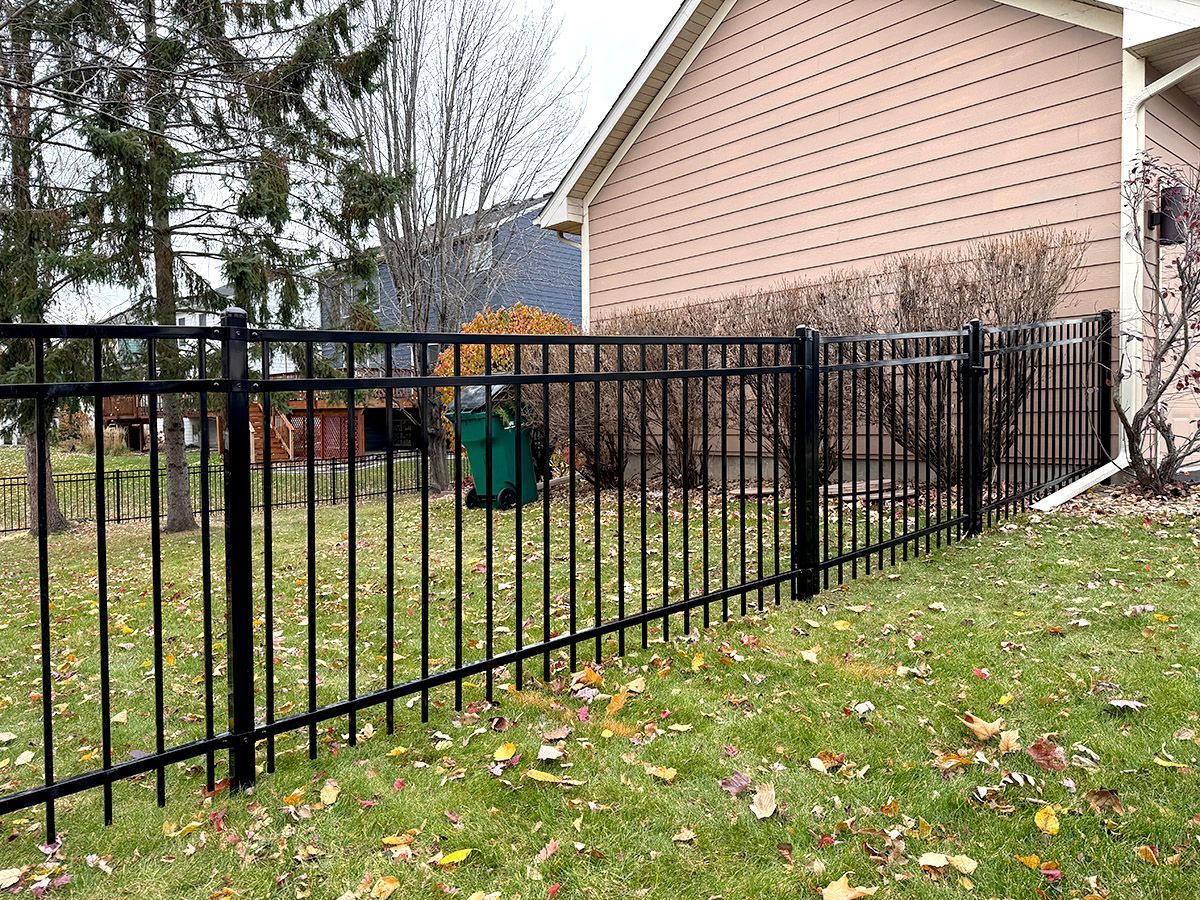 A black metal fence runs along the side of a beige house, bordering a lawn with fallen leaves on a cloudy day.