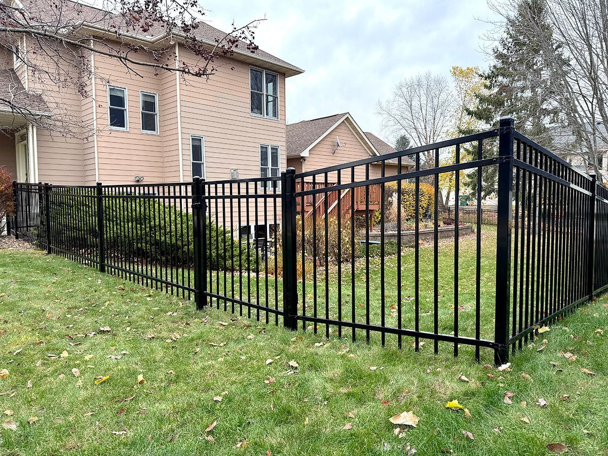 A black metal fence stretches across a suburban backyard with a multi-story, light-colored house in the background.