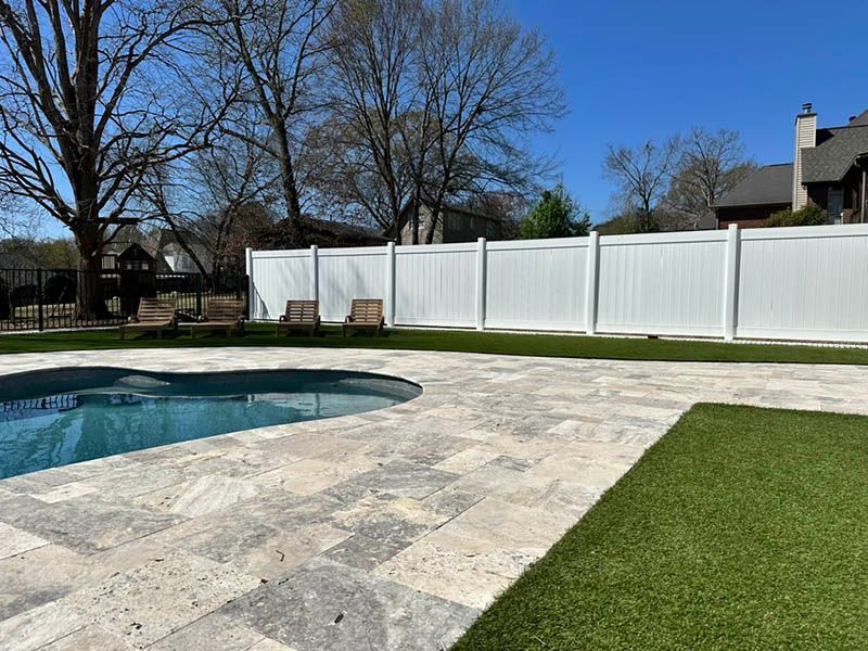 Backyard patio with a pool, stone paving, green lawn, and white privacy fence under a blue sky