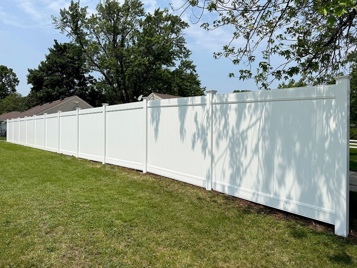 A long, white, solid vinyl privacy fence standing in a grassy yard under a bright, sunny sky.